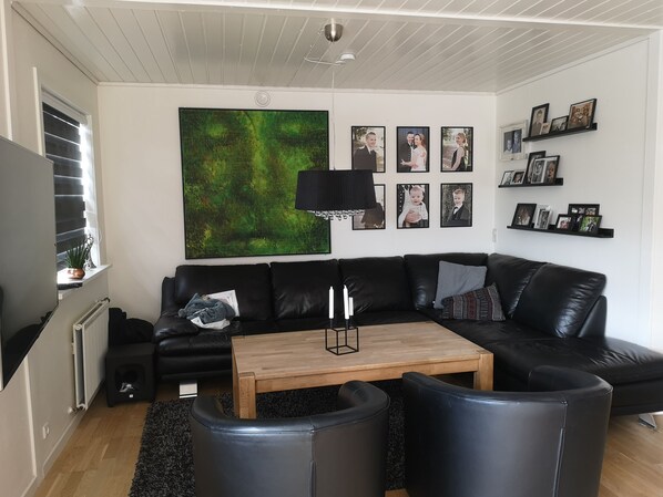 Un salon moderne avec un canapé en cuir noir, une table basse en bois et des fauteuils en cuir noir. Un grand tableau abstrait orne le mur, ainsi qu’un lampadaire noir et une étagère où sont exposées des photos encadrées.