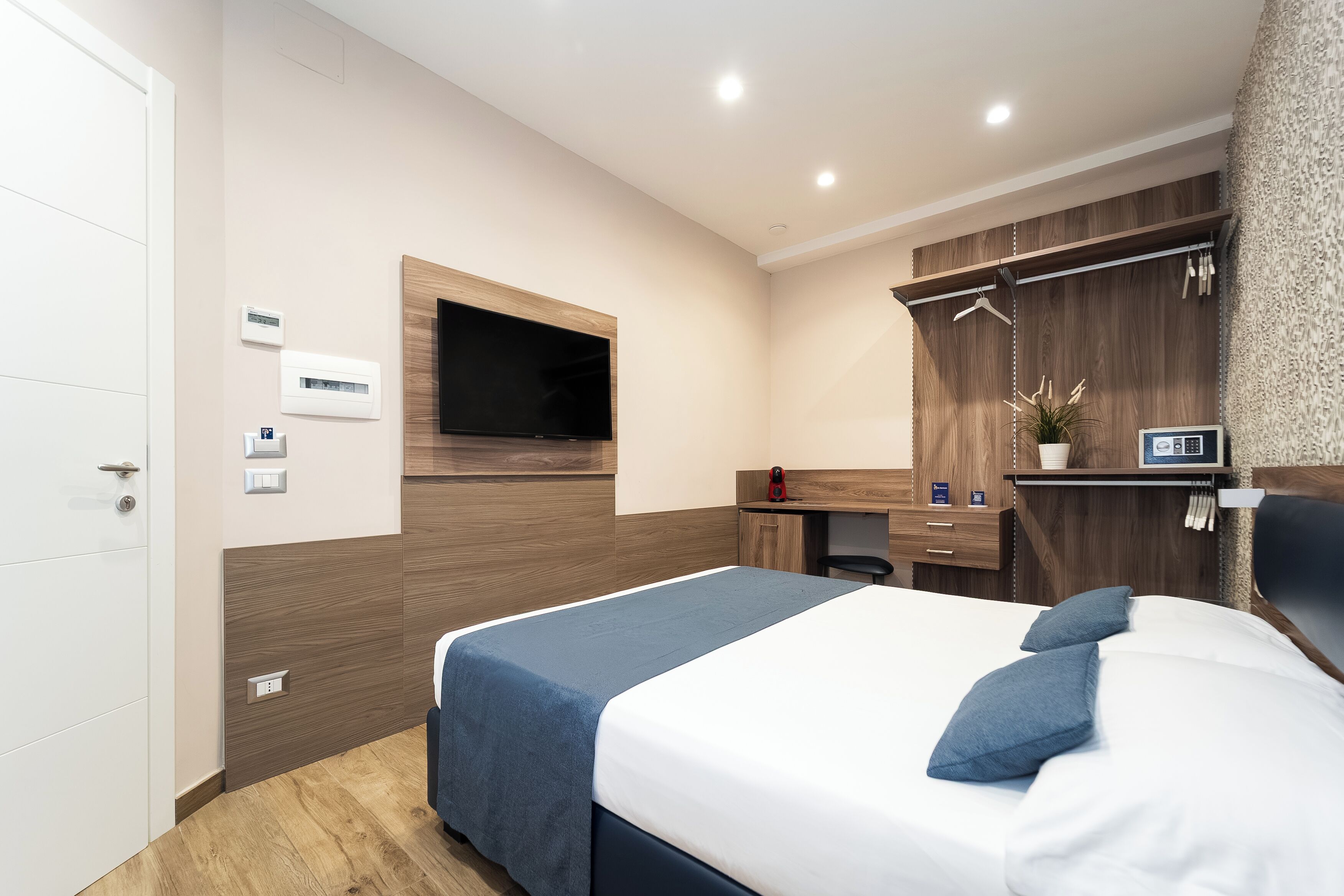 A modern hotel room with a bed, a TV mounted on the wall, a desk, and a wardrobe.
