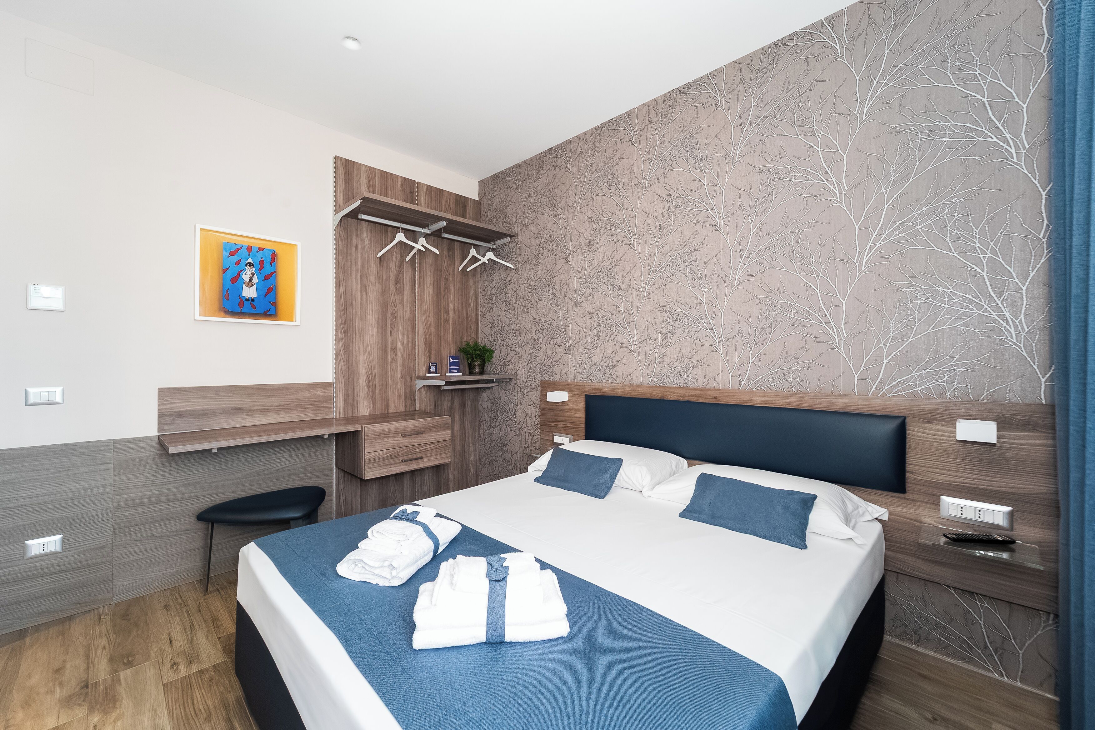 A modern hotel room with a large bed, a bench, a wall-mounted TV, and a small shelf with a plant.