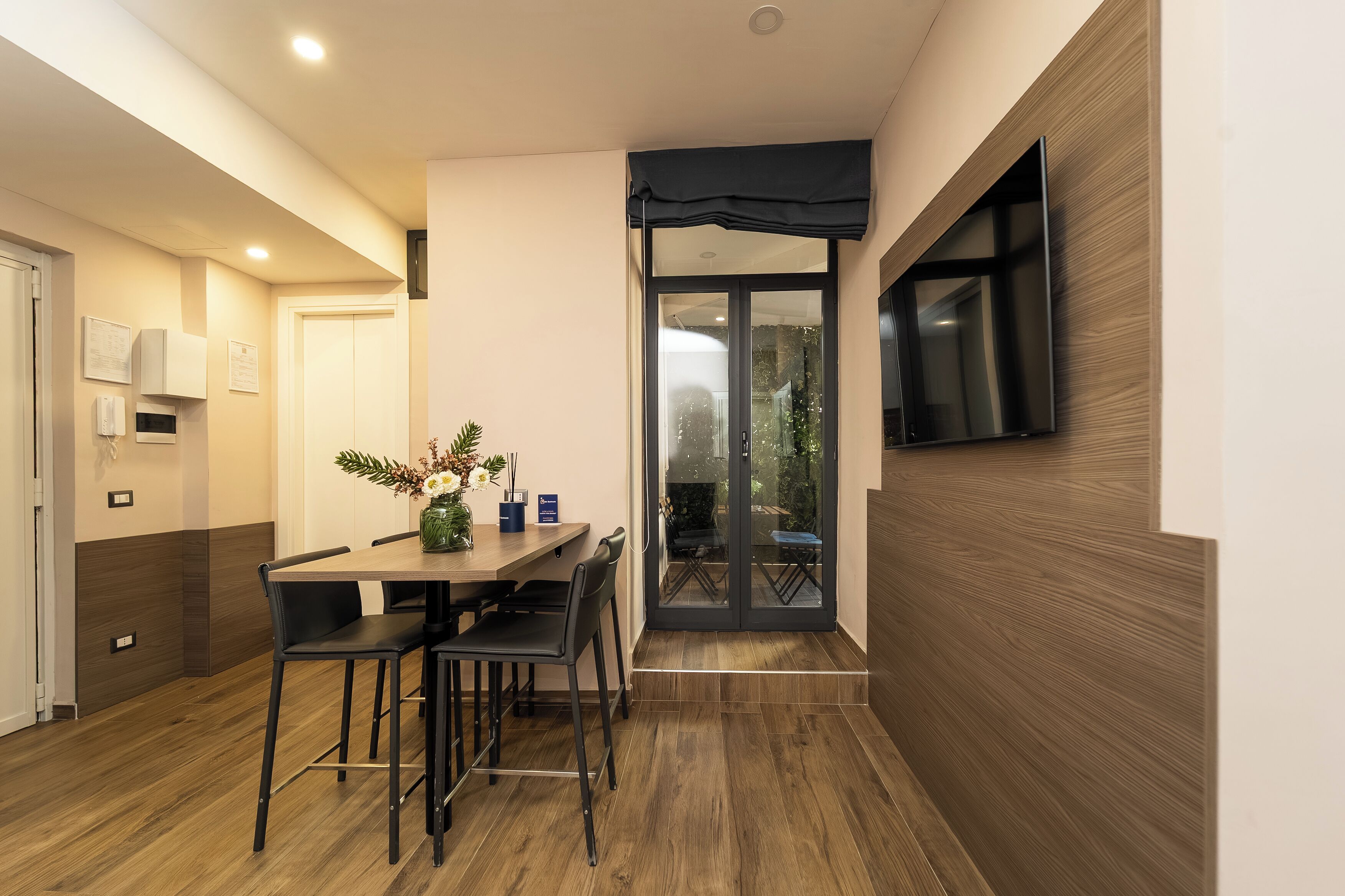 A modern dining area with a wooden table and chairs, a TV mounted on the wall, and a sliding glass door leading to an outdoor area.