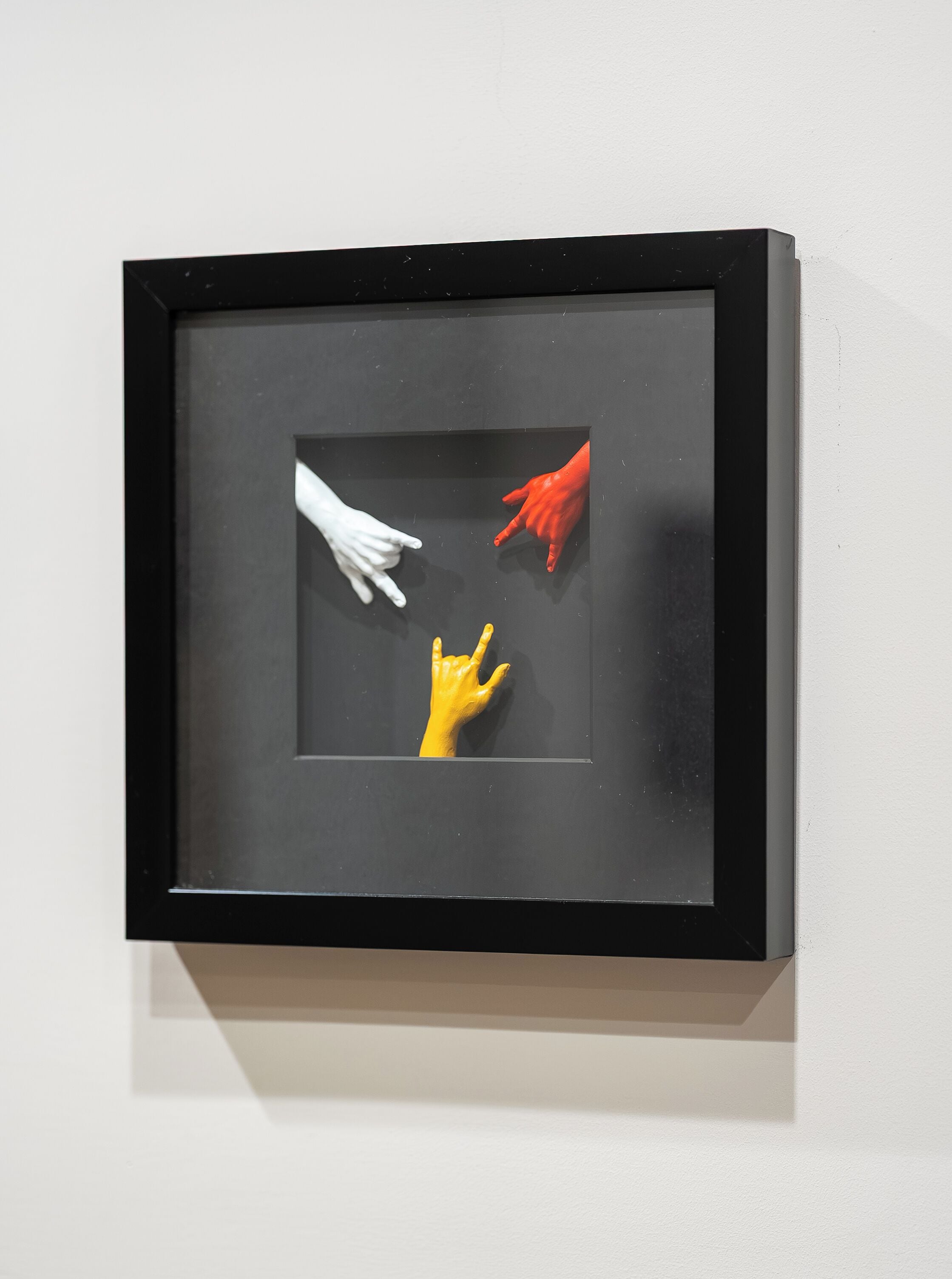 A framed artwork with three hands in different colors on a white wall.