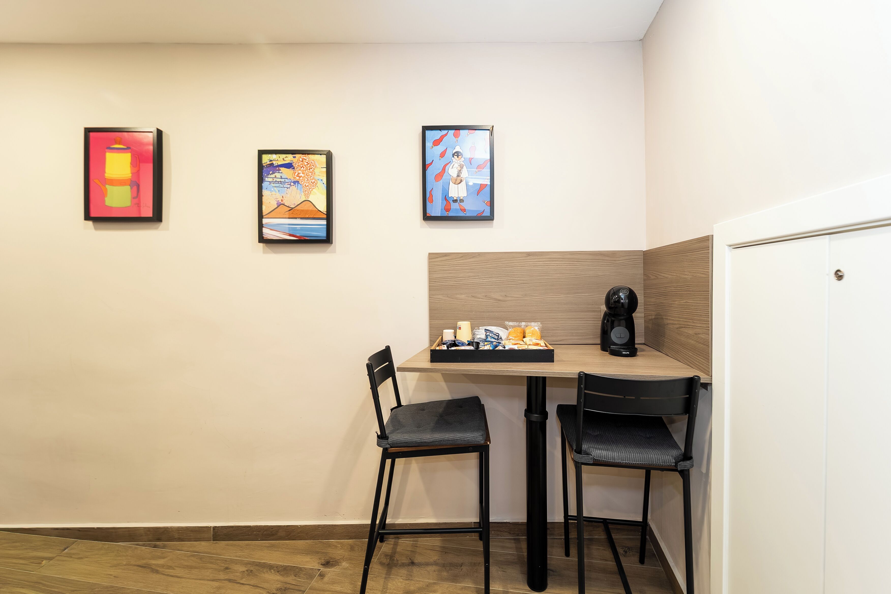 A small, modern room with a wooden table, two black chairs, and a coffee machine. There are three framed artworks on the wall.