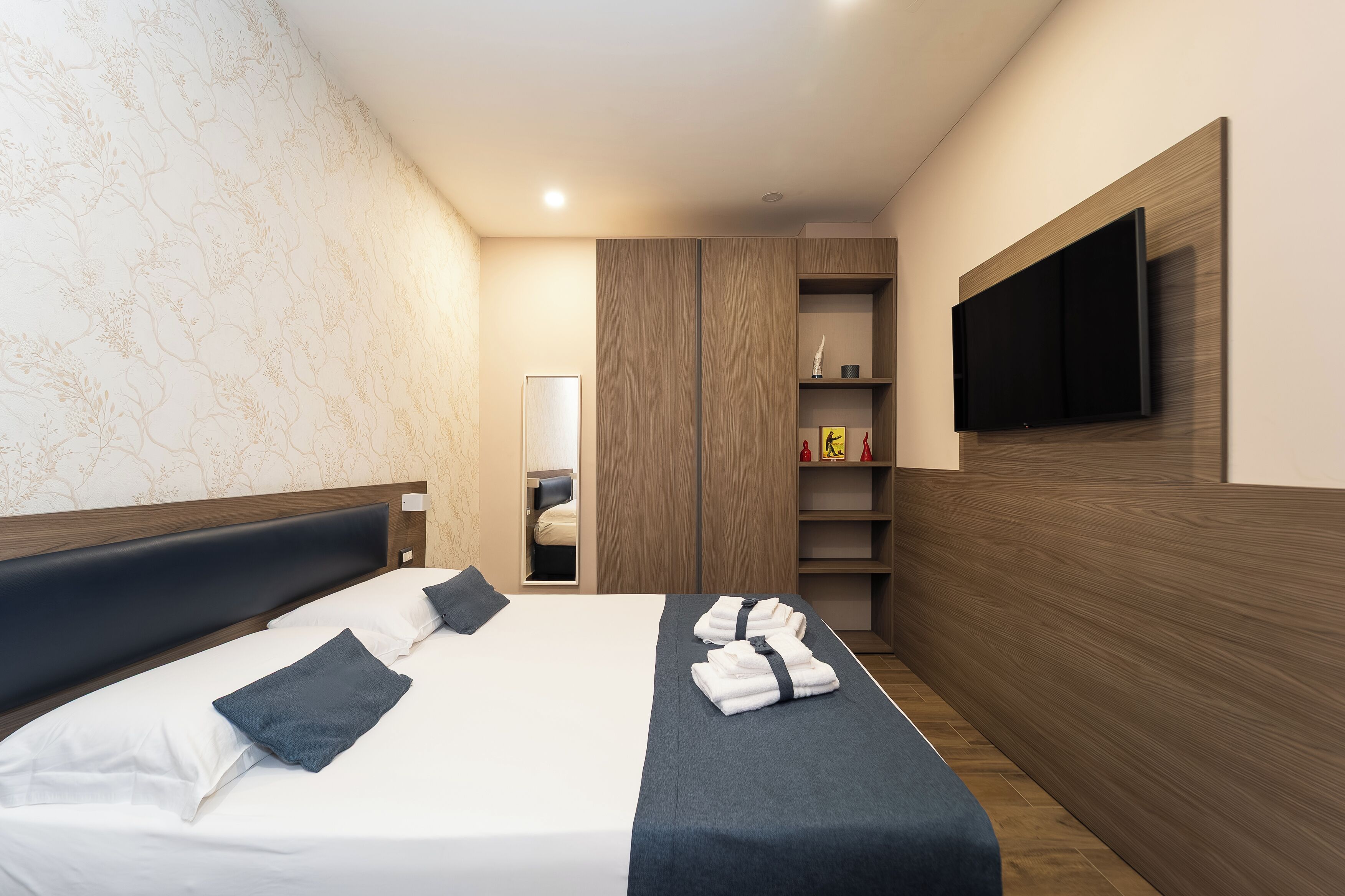 A hotel room with a bed, a TV mounted on the wall, a wardrobe, and a small shelf with items.
