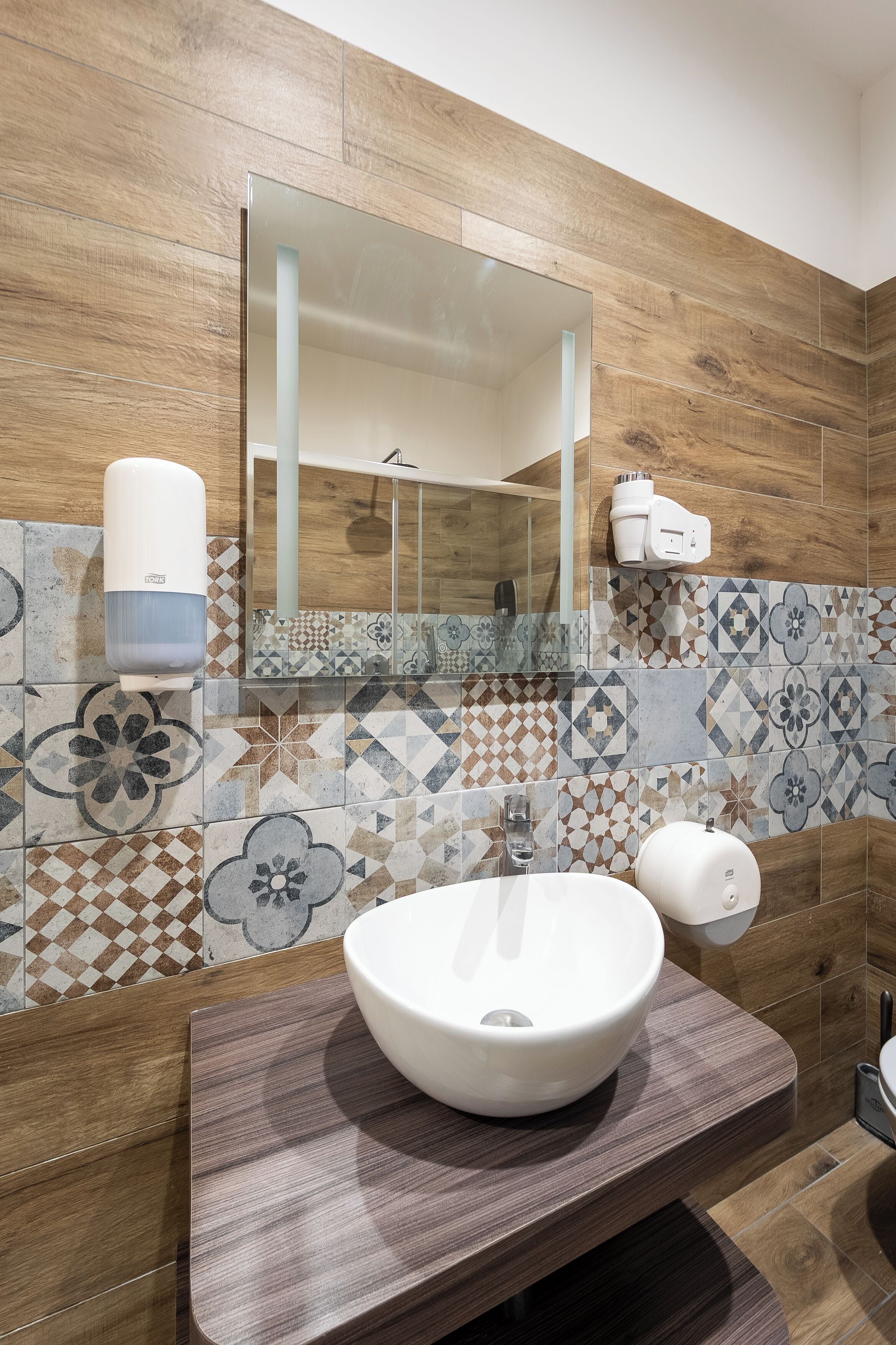 A bathroom with a wooden wall, a tiled backsplash, a white sink, a mirror, and a towel dispenser.