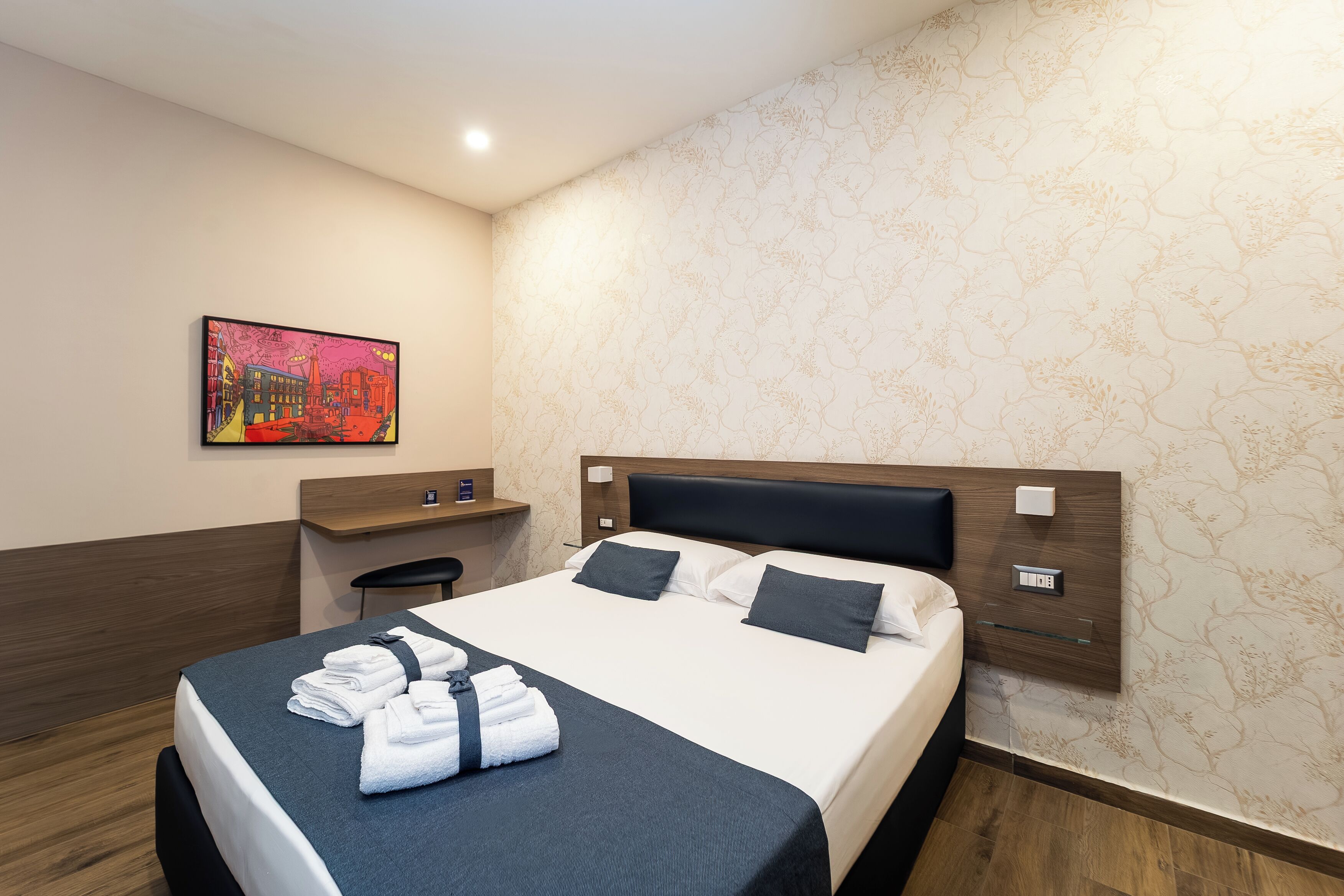 A hotel room with a bed, a desk, and a painting on the wall.