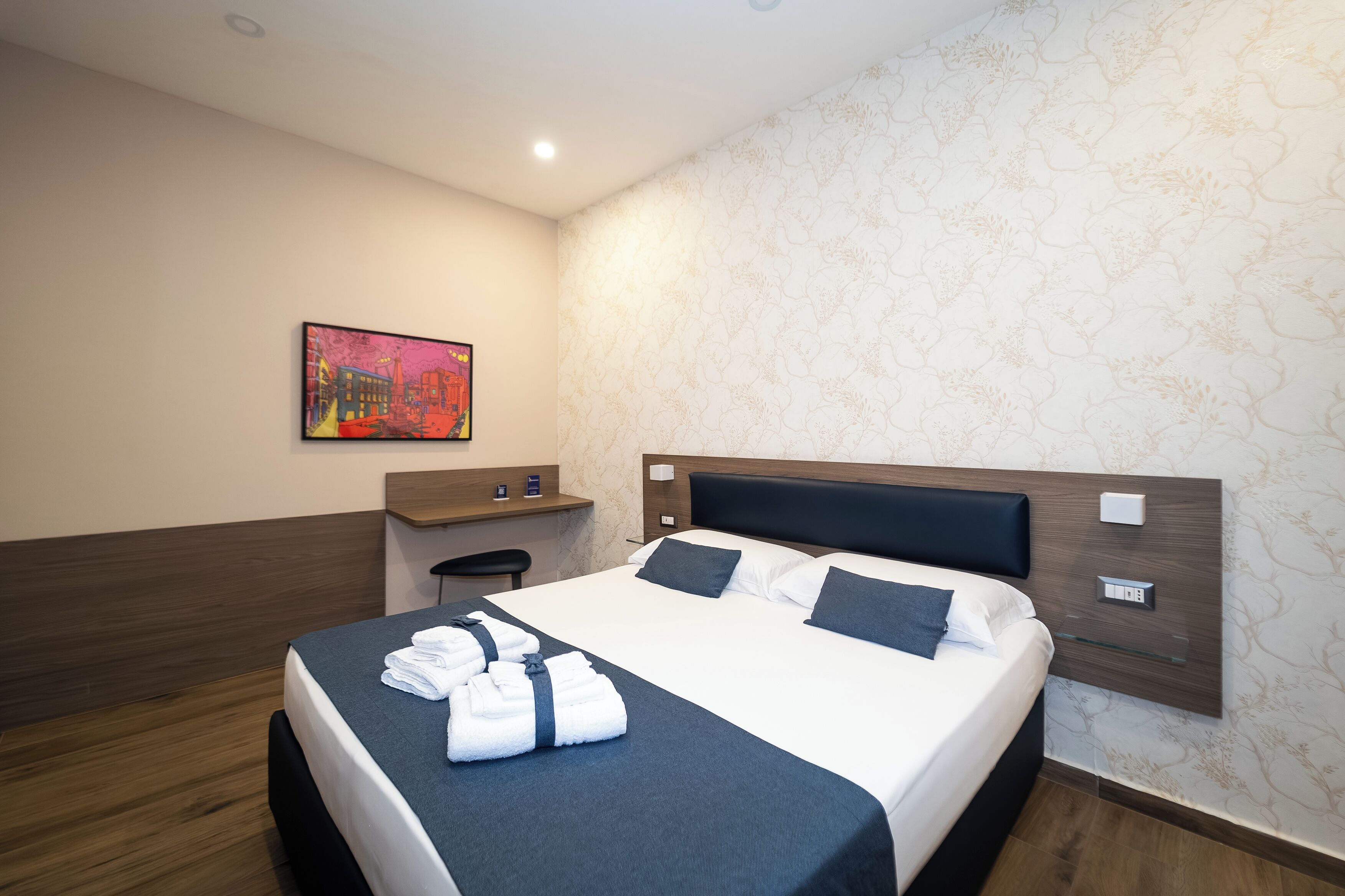 Una habitación de hotel con una cama, un escritorio, una silla, un cuadro en la pared y un papel tapiz estampado.