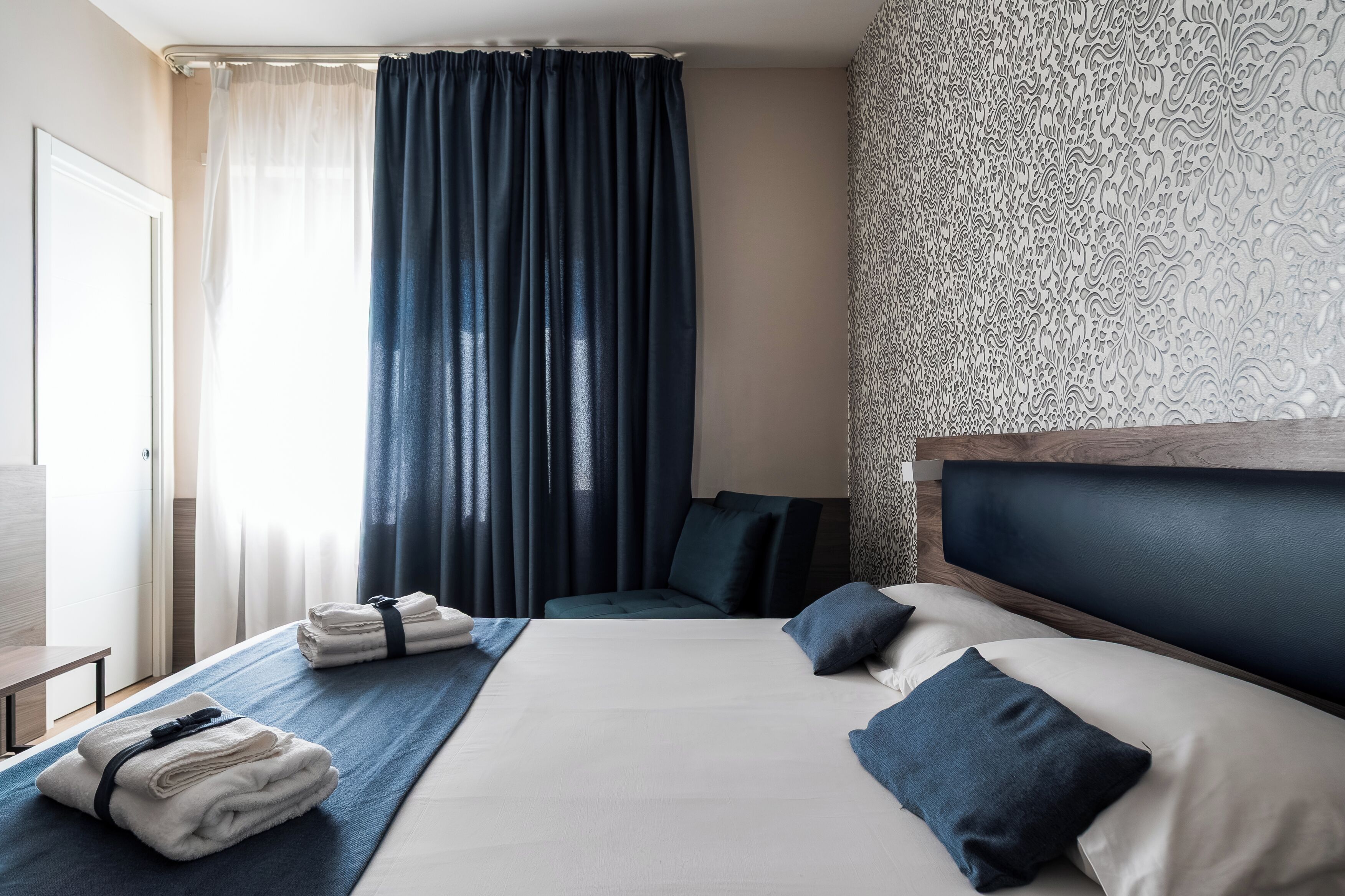 A hotel room with a bed, pillows, a blue blanket, and a set of towels.