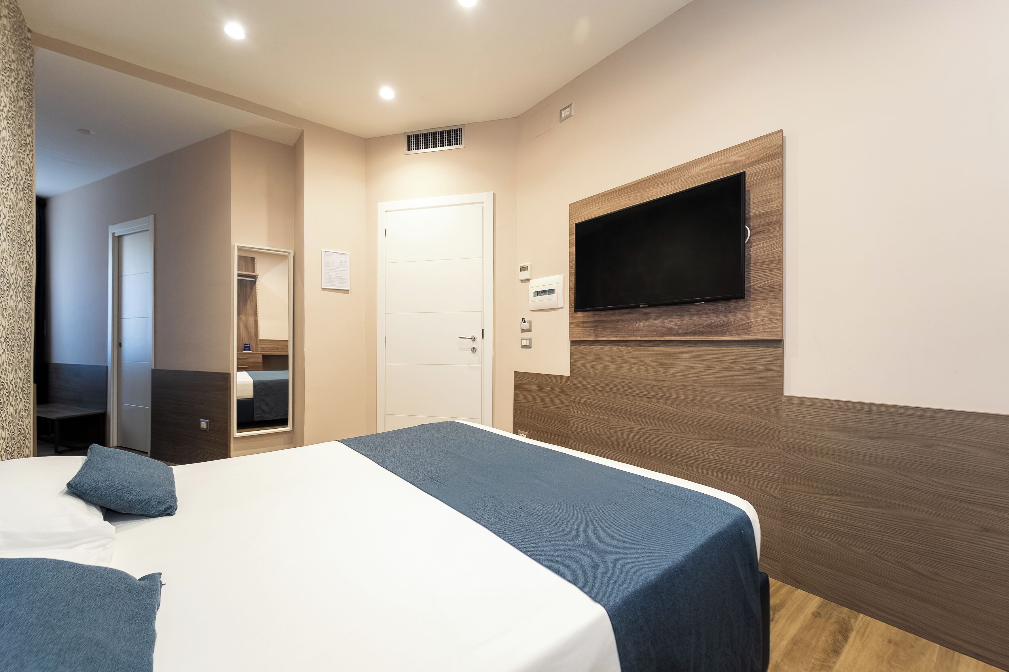 Ett hotellrum med en säng, en tv som är monterad på väggen, en dörr och ett litet fönster.