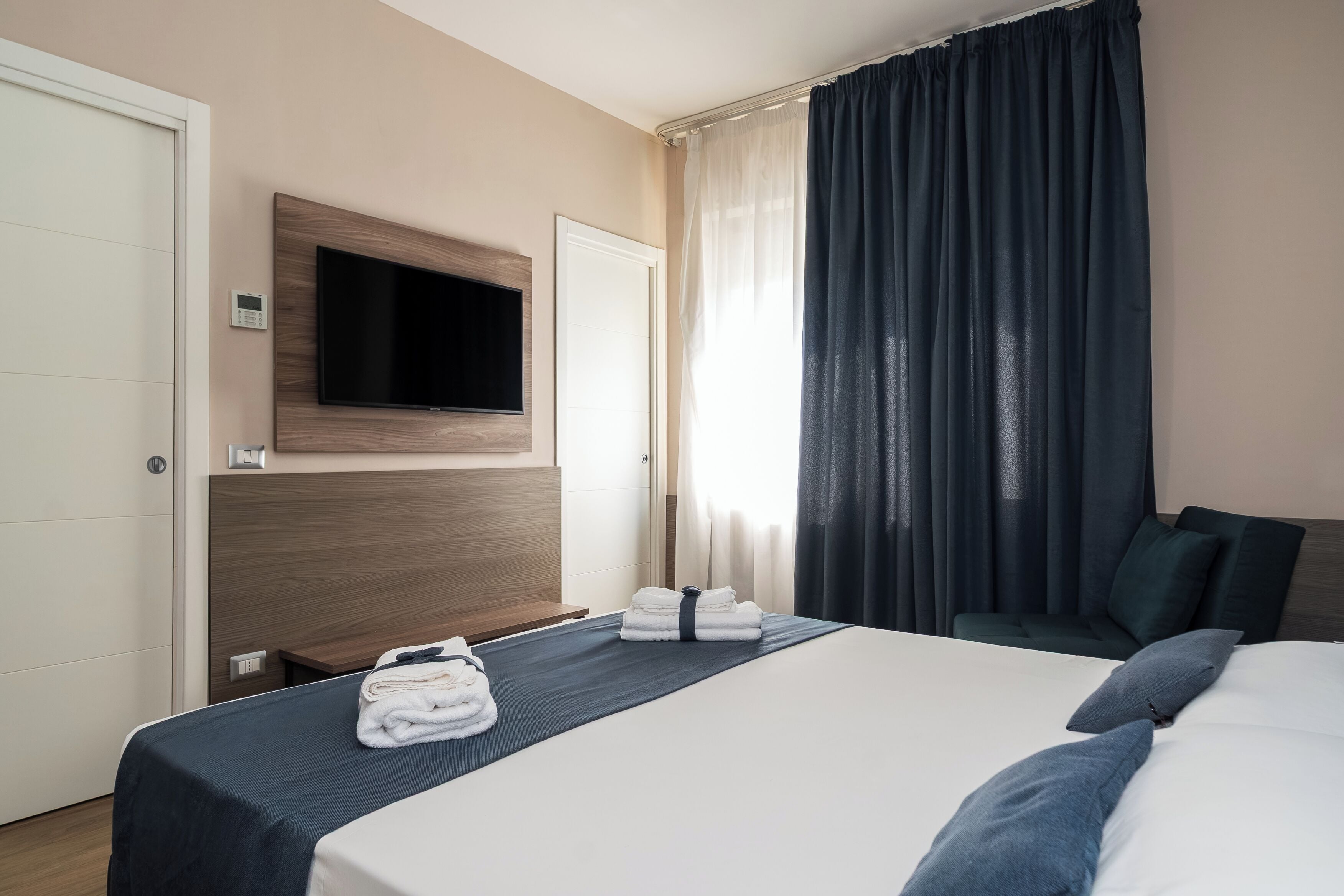 Una camera d'albergo con un letto, una televisione a parete, una finestra con tende e una porta.
