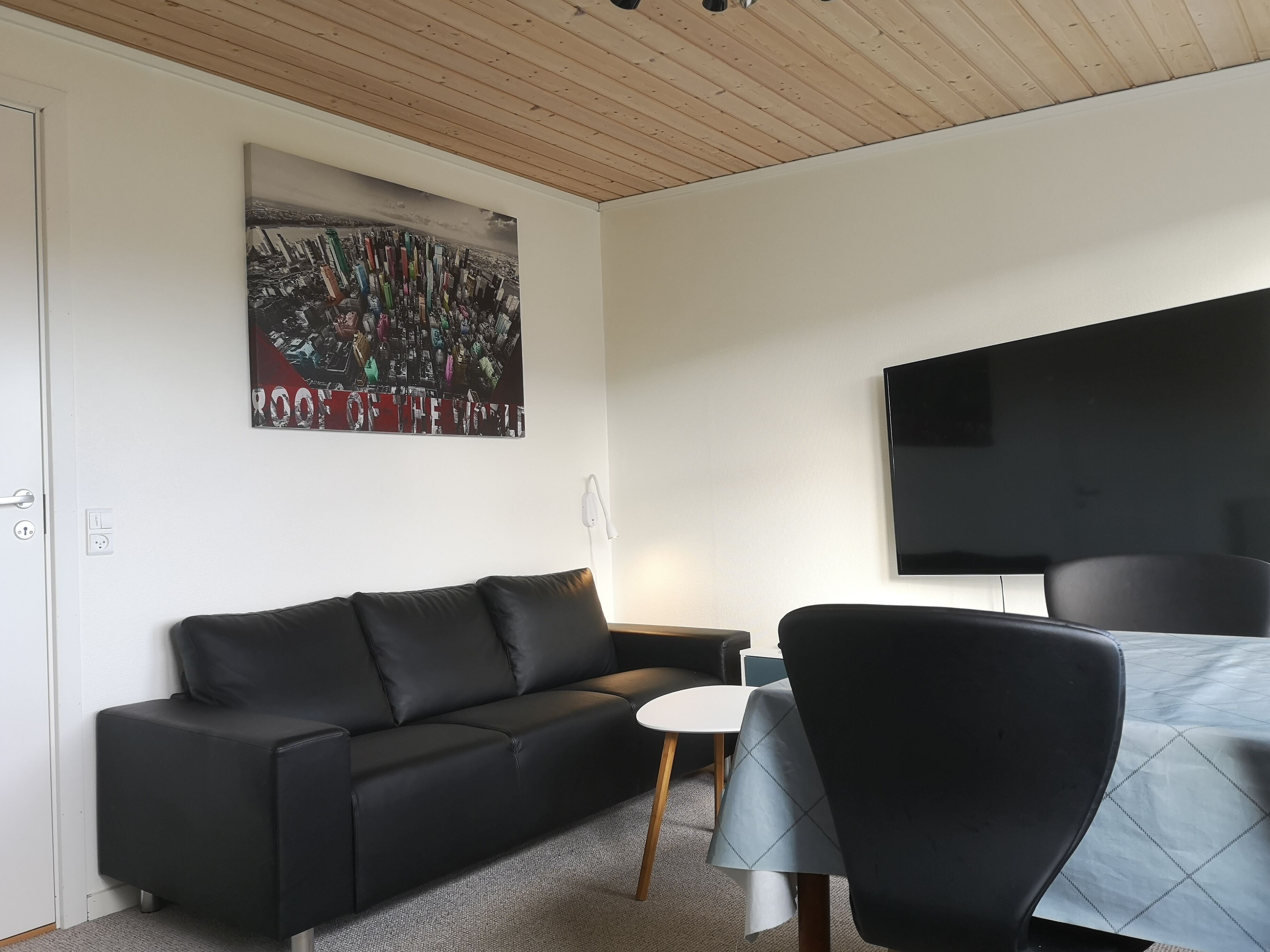 A modern living room with a black leather sofa, a small round table with a white tablecloth, and a large flat-screen TV mounted on the wall.
