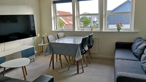 Una sala de estar moderna con televisor, mesa de comedor, sillas, sofá y ventanas amplias con vistas a edificios y árboles.