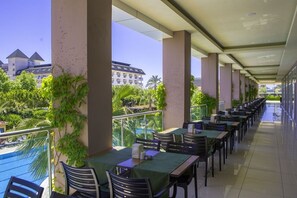 A covered outdoor dining area with tables and chairs, a pool in the background, and a view of a hotel building. - Telatiye Resort Hotel (Alanya)