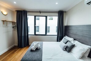 A modern hotel room with a large bed, dark curtains, a small table with towels, and a window with a view of buildings. - UNO Hotel Sydney (Sydney)