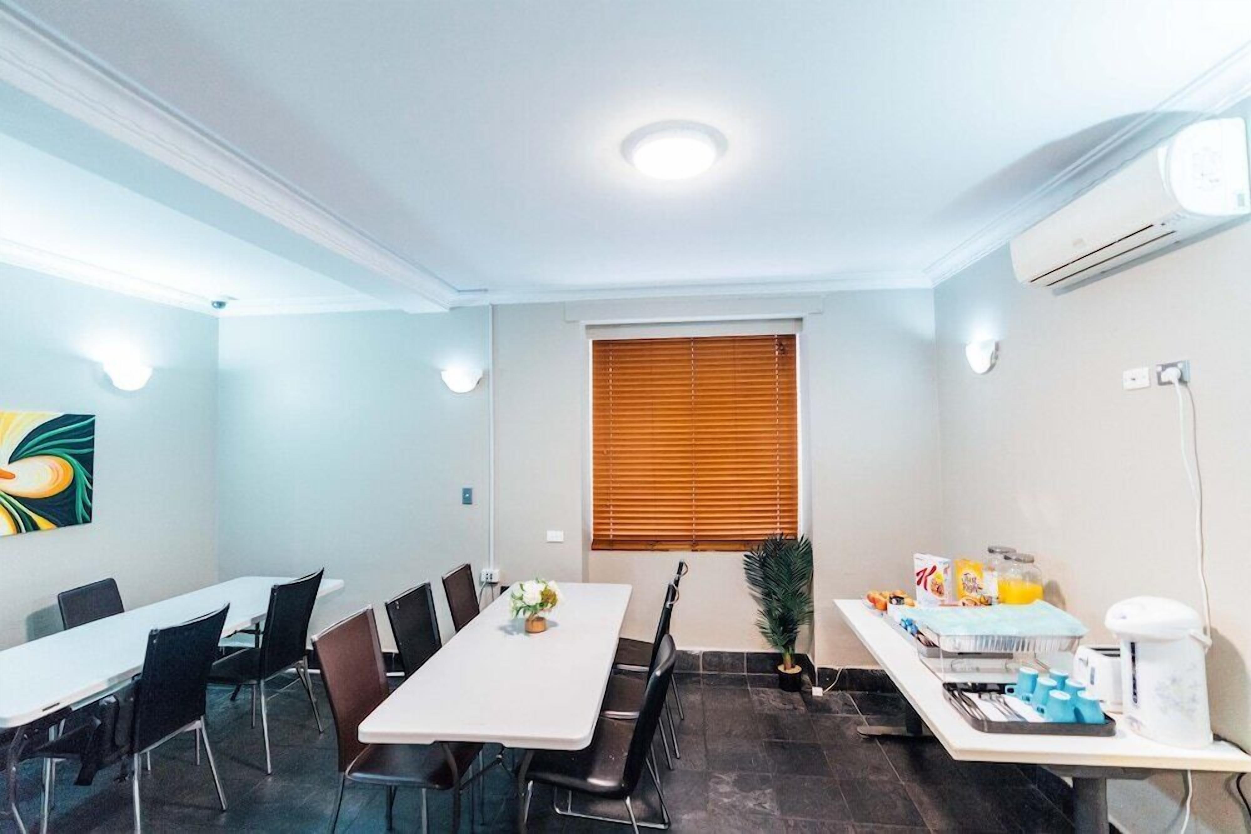 Una sala de conferències amb una taula llarga, cadires, una finestra amb persianes, una planta en test i una taula de buffet amb aliments i begudes.
