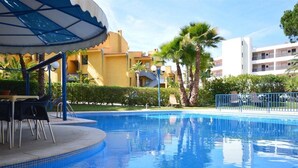 A swimming pool with a sunshade, palm trees, and a building in the background. - Apartments Rio By Garvetur (Loulé)