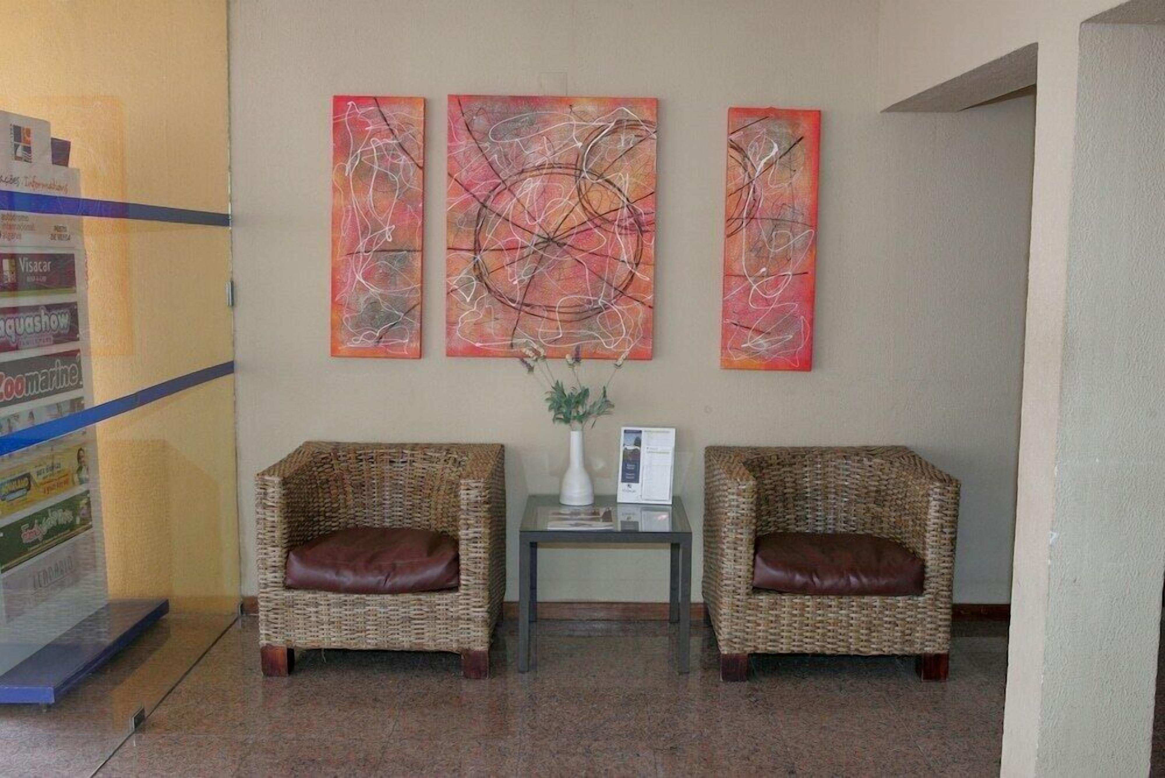 A lobby with two wicker armchairs, a small table with a vase and a brochure, and three abstract paintings on the wall.