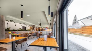 A modern restaurant with wooden tables and chairs, pendant lighting, and a view of the outside. - Pension Alpenrose (Kaprun)