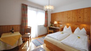 A hotel room with a wooden headboard, a bed with white linens, a desk with a lamp, a chair, and a table with a bottle and glasses. - Pension Alpenrose (Kaprun)