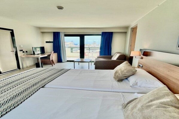 A hotel room with a bed, a desk with a computer, a chair, and a view of the city through the window.