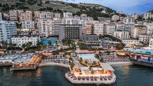 Paisatge urbà costaner amb edificis alts, una marina amb vaixells i una zona de platja amb ombrel·les i gandules.