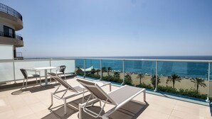 Un balcón con sillones de descanso y una mesa con vista a una playa con palmeras y el océano.