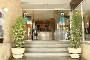 L’entrée d’un hôtel, avec un escalier menant au hall d’accueil, des plantes en pot de chaque côté et des panneaux indiquant les équipements disponibles, comme un minibar et un restaurant.