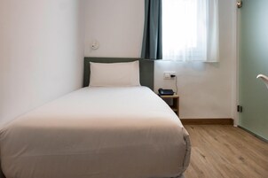 Una cama individual con colchón y almohada blancos, una mesita de noche con un teléfono y un enchufe de pared.