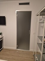 Una habitación con una puerta cerrada, un televisor montado, un estante blanco y una planta en maceta.