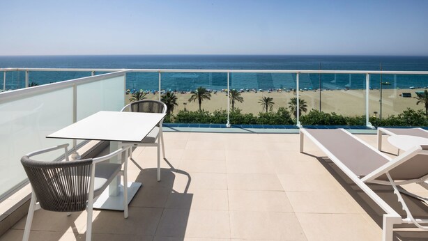 A balcony with a table and chairs, overlooking a beach with palm trees and the ocean. - Fragata Hotel (Calella)