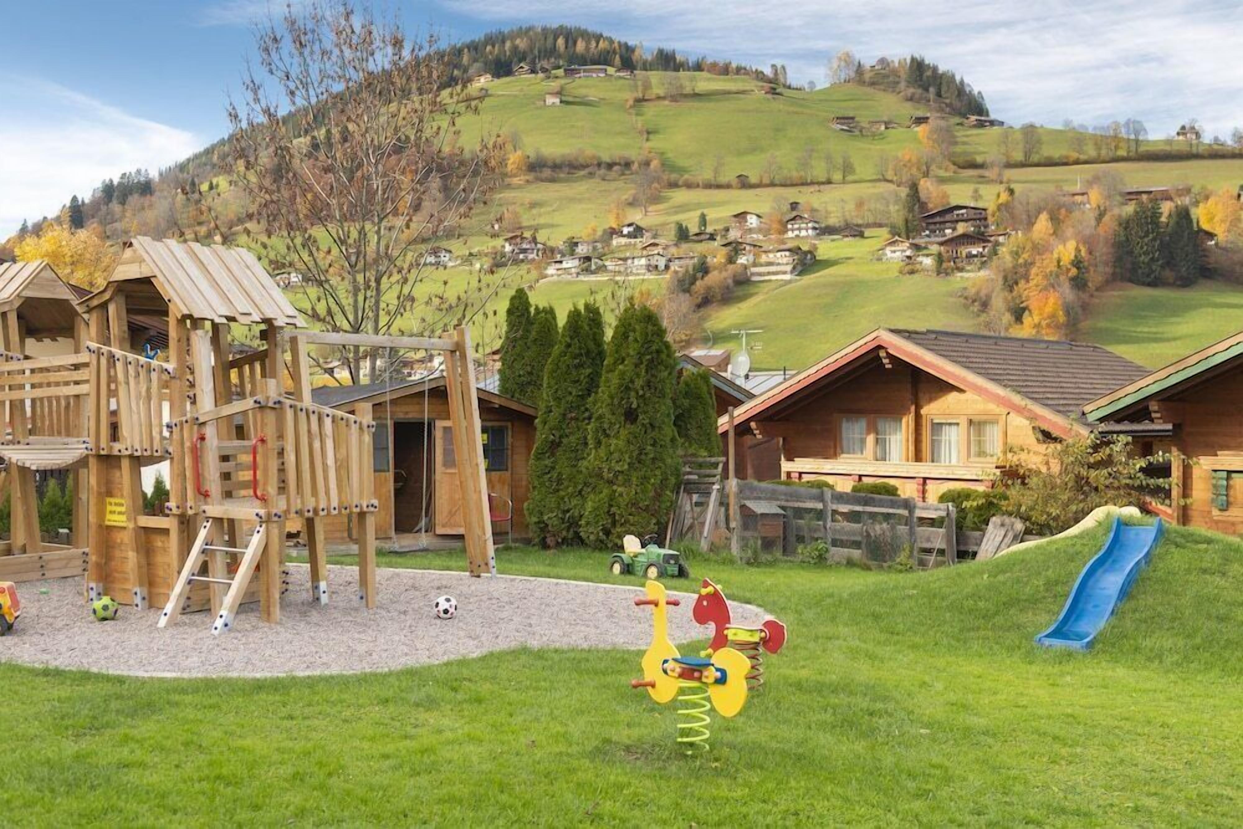A playground with a wooden climbing structure, a slide, and a swing set in a rural village setting.