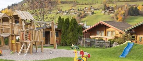 A playground with a wooden climbing structure, a slide, and a swing set in a rural village setting.