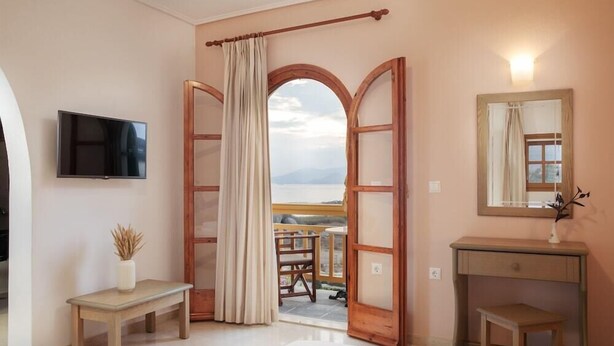 A room with a large arched doorway leading to a balcony with a view, a flat-screen TV mounted on the wall, a wooden bench, and a small table with a vase. - Proteas Studios (Naxos)