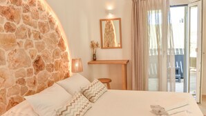 A bedroom with a stone wall, a bed with white bedding, a wooden nightstand, a mirror, and a view of the outdoors. - Proteas Studios (Naxos)