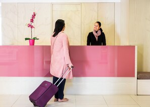 Un espace d’accueil avec un mur recouvert de panneaux en bois, un comptoir d’accueil rose et une valise violette posée sur le sol.