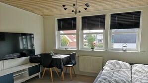 A modern living room with a flat-screen TV, a dining table with chairs, a sofa, and a window with blinds. - Rose Cottage 15m2 Tórshavn with Parking (Hoyvík)