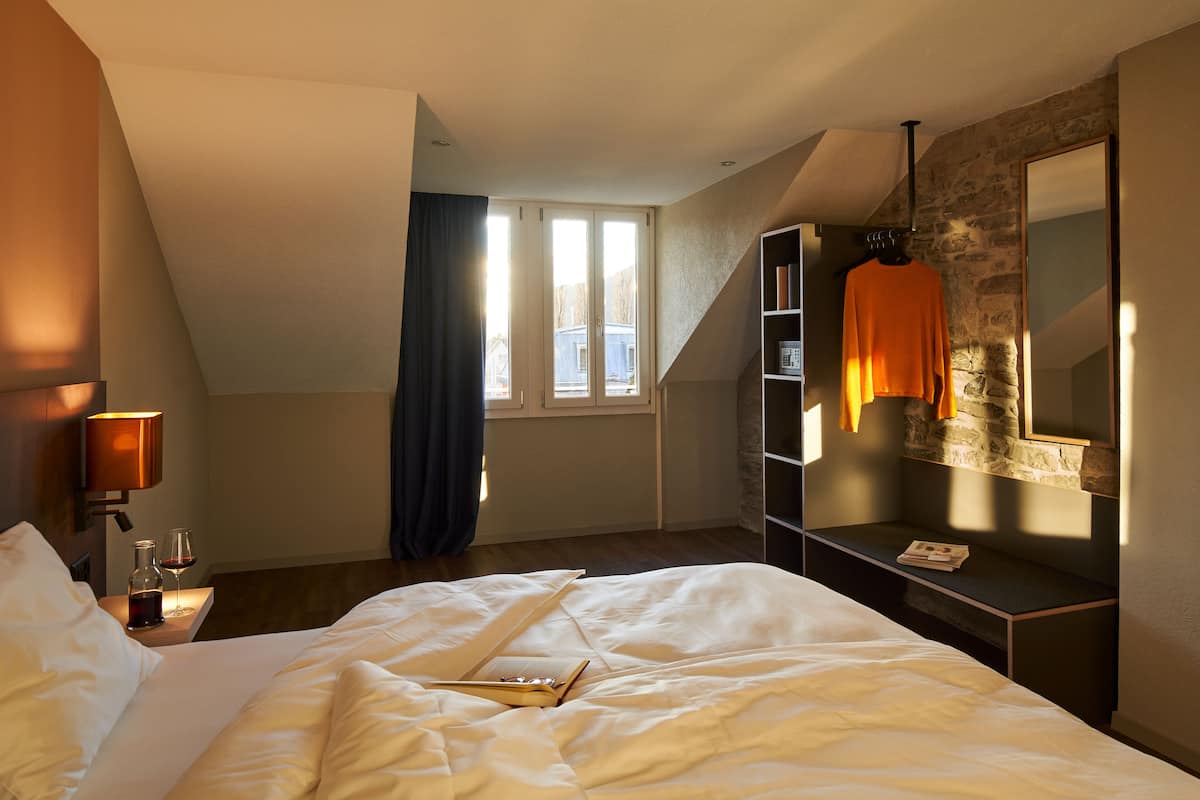 A bedroom with a bed, a bedside table with a lamp, a window with curtains, a stone wall, and a hanging garment.