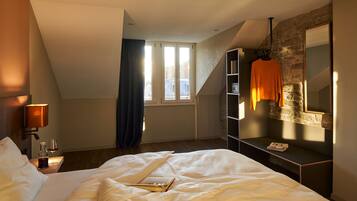 Un dormitorio con cama, mesita de noche con lámpara, ventana con cortinas, pared de piedra y ropa colgada.