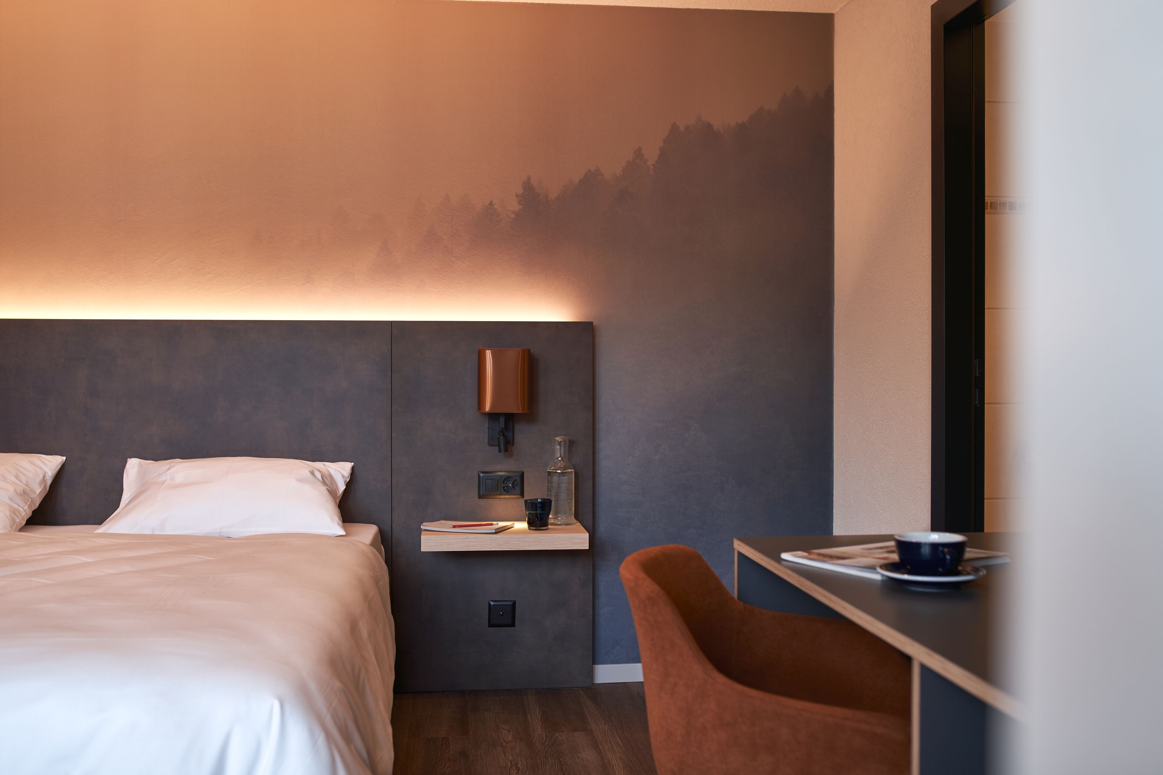 Una camera d'albergo moderna con un letto, un comodino, una scrivania con sedia e una lampada a parete.