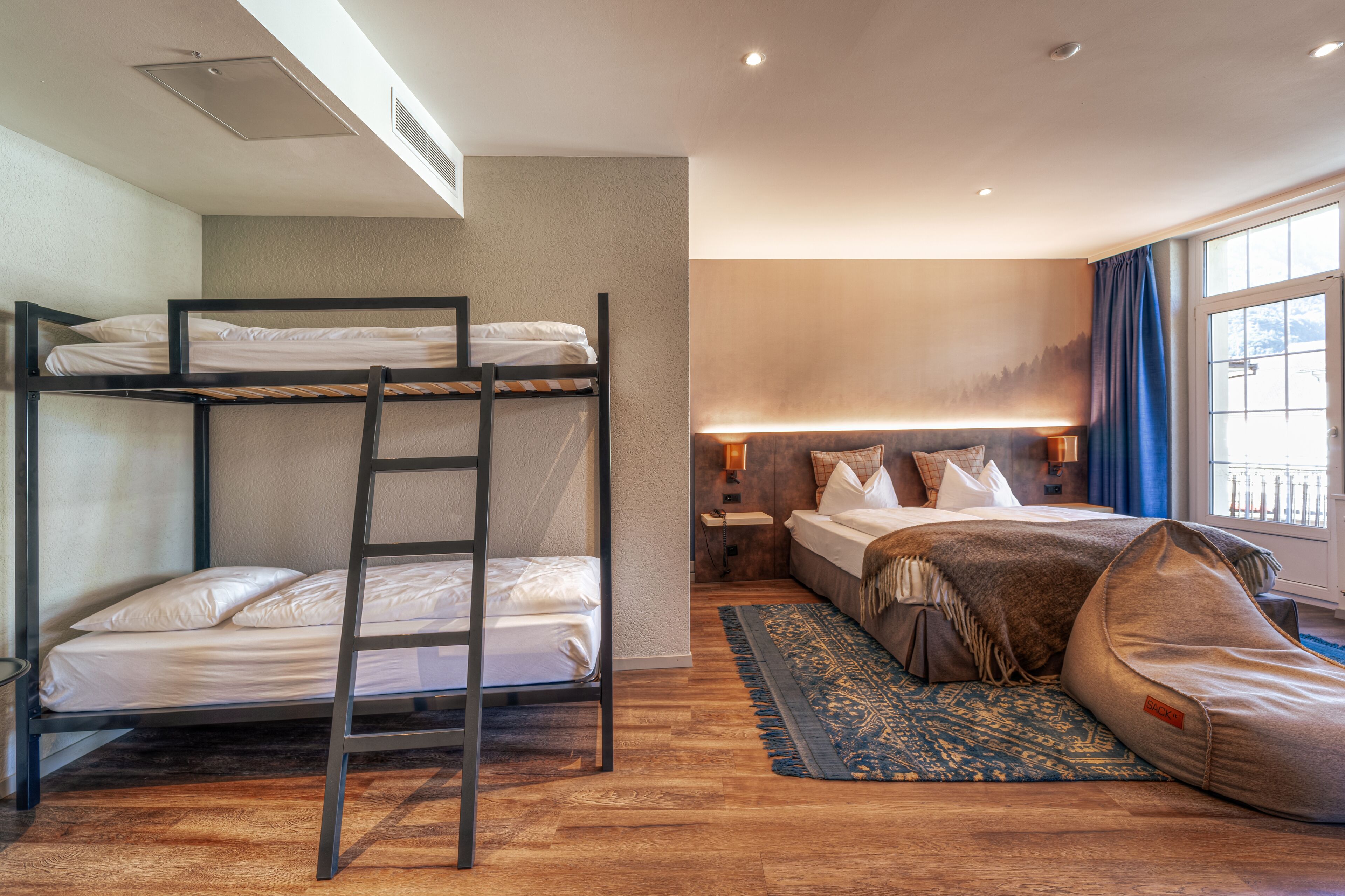 A hotel room with bunk beds, a separate bed, a bedside table, a lamp, a window with curtains, and a patterned rug.
