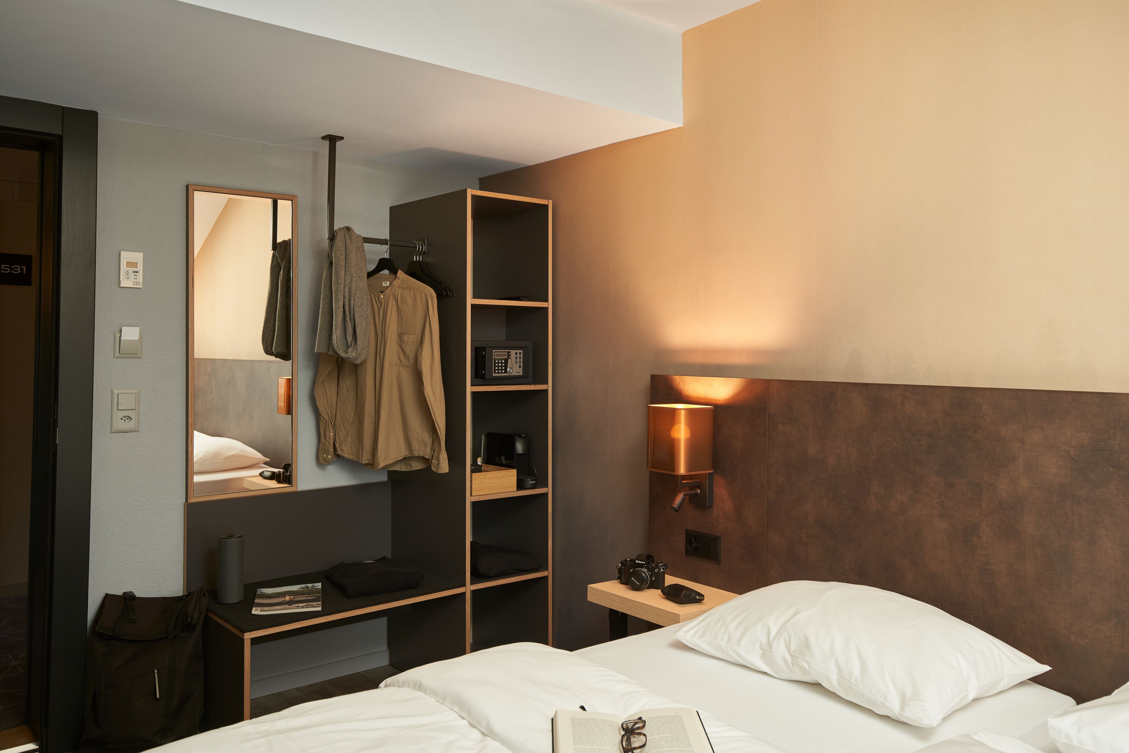 A hotel room with a bed, a bedside table, a wardrobe, a mirror, and a wall-mounted lamp.