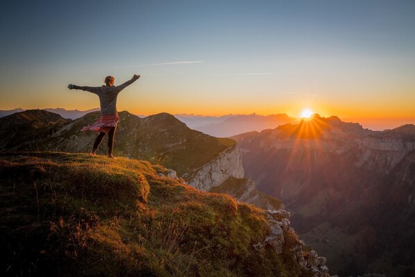 Una persona de pie en una colina cubierta de hierba, con los brazos extendidos, contemplando un paisaje montañoso al atardecer.