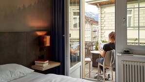 Habitación de hotel con cama, mesita de noche con libros, ventana con vistas a edificios y montañas, y balcón con mesa y silla.