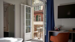 Habitación de hotel con cama, escritorio con silla, televisor y puerta corrediza de vidrio que da a un balcón exterior con mesa y sillas.