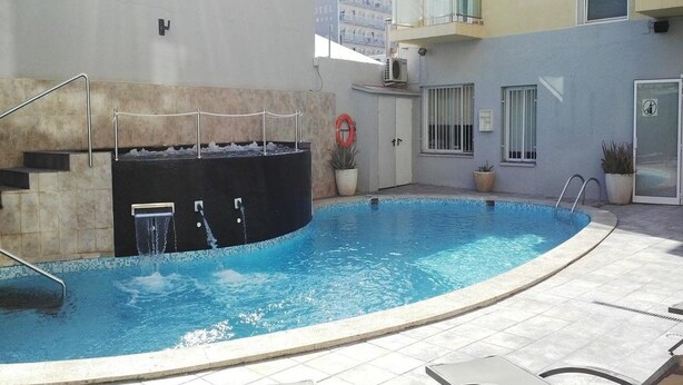 A swimming pool with a water feature, surrounded by a tiled patio and a building with balconies. - Sun Beach (Lloret de Mar)