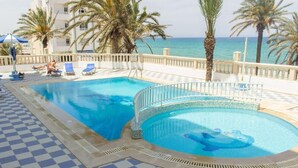Zona de piscina con dos piscinas, sillas de descanso, palmeras y vista al mar.