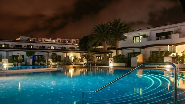 A swimming pool with a modern design, surrounded by lounge chairs and umbrellas, with a building in the background. - Relaxia Lanzaplaya (Tías)