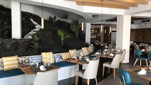 A modern restaurant with a stone wall and plants, wooden ceiling beams, and a variety of seating options. - Relaxia Lanzaplaya (Tías)