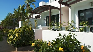A modern building with a white facade, a covered outdoor seating area, and lush greenery including banana trees and flowering plants. - Relaxia Lanzaplaya (Tías)