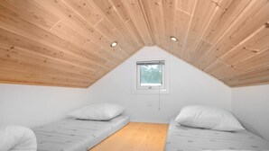 A small, single-room attic bedroom with wooden ceiling beams, two single beds, a window, and a wooden floor.