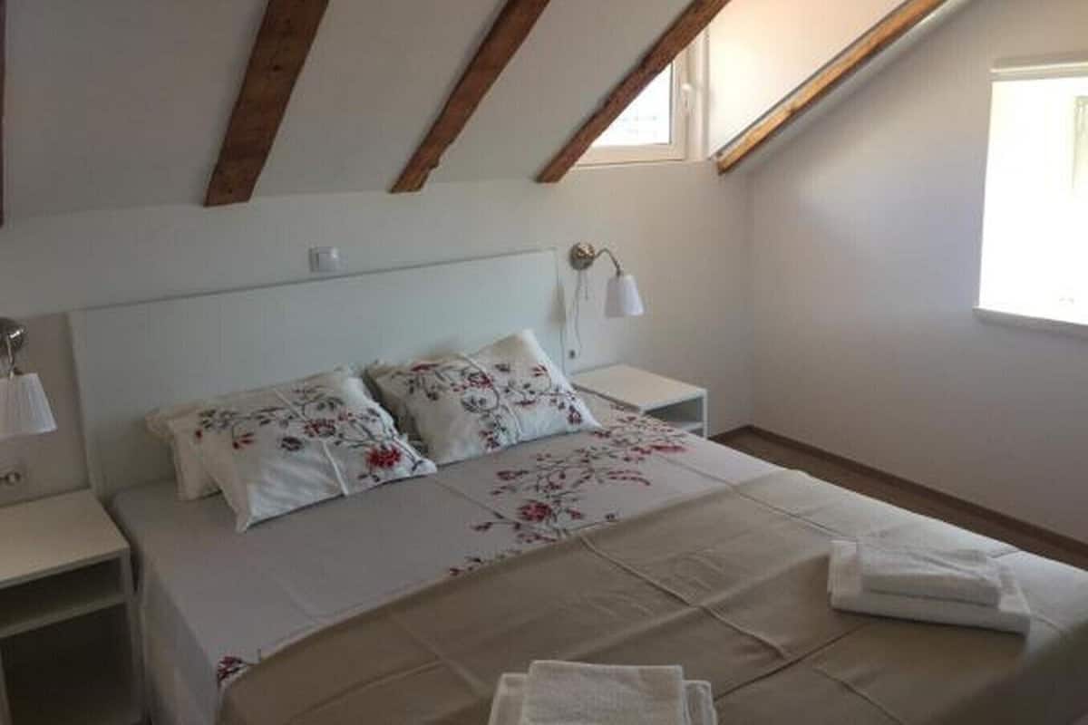A bedroom with a sloped ceiling, a bed with floral bedding, a bedside table with a lamp, and a window providing natural light.
